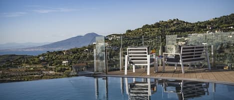 An infinity pool with clear water reflecting the surroundings, two white lounge chairs, and a view of a mountainous landscape in the background.