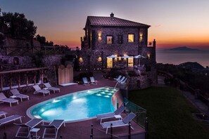 A stone building with a swimming pool, lounge chairs, and a view of the sea at sunset.