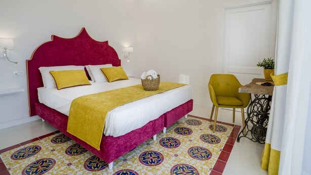 Una cama con cabecera roja, una manta amarilla y dos almohadas. Junto a la cama, una pequeña mesa con una silla.