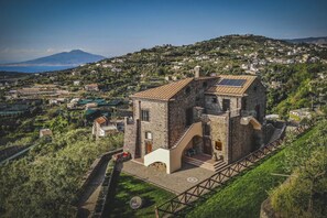 A stone building with a staircase leading to a terrace, surrounded by greenery and a mountainous backdrop.