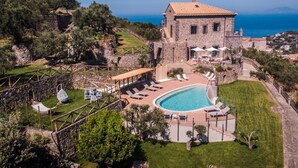 Un edificio de piedra con piscina y mobiliario exterior, rodeado de vegetación y vistas a un cuerpo de agua.