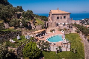 A stone building with a pool and outdoor seating, surrounded by greenery and overlooking a body of water. - MyExclusiveItaly - Villa Breathtaking view near Nerano (Massa Lubrense)
