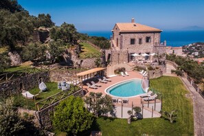 Un edificio de piedra con piscina y mobiliario exterior, rodeado de vegetación y vistas a un cuerpo de agua.