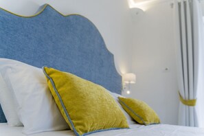 Una cama con cabecera azul, almohadas blancas y amarillas, y una pared blanca con una lámpara.