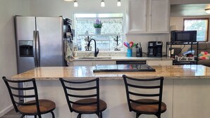 Una cocina con una isla central, taburetes de bar, refrigerador, cafetera y una ventana con vistas.