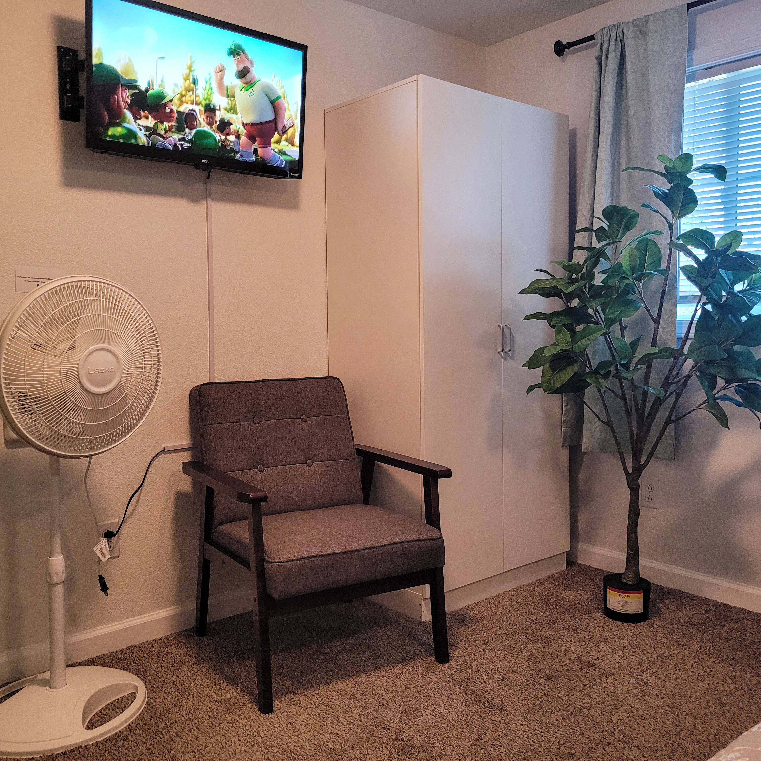 A room with a TV mounted on the wall, a standing fan, a chair, a wardrobe, and a potted plant near a window.