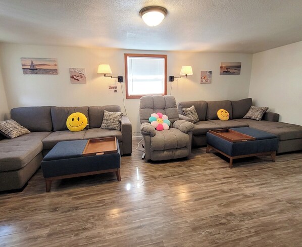Una sala de estar con un sofá seccional, dos otomanas, un sillón reclinable y una ventana con persianas.