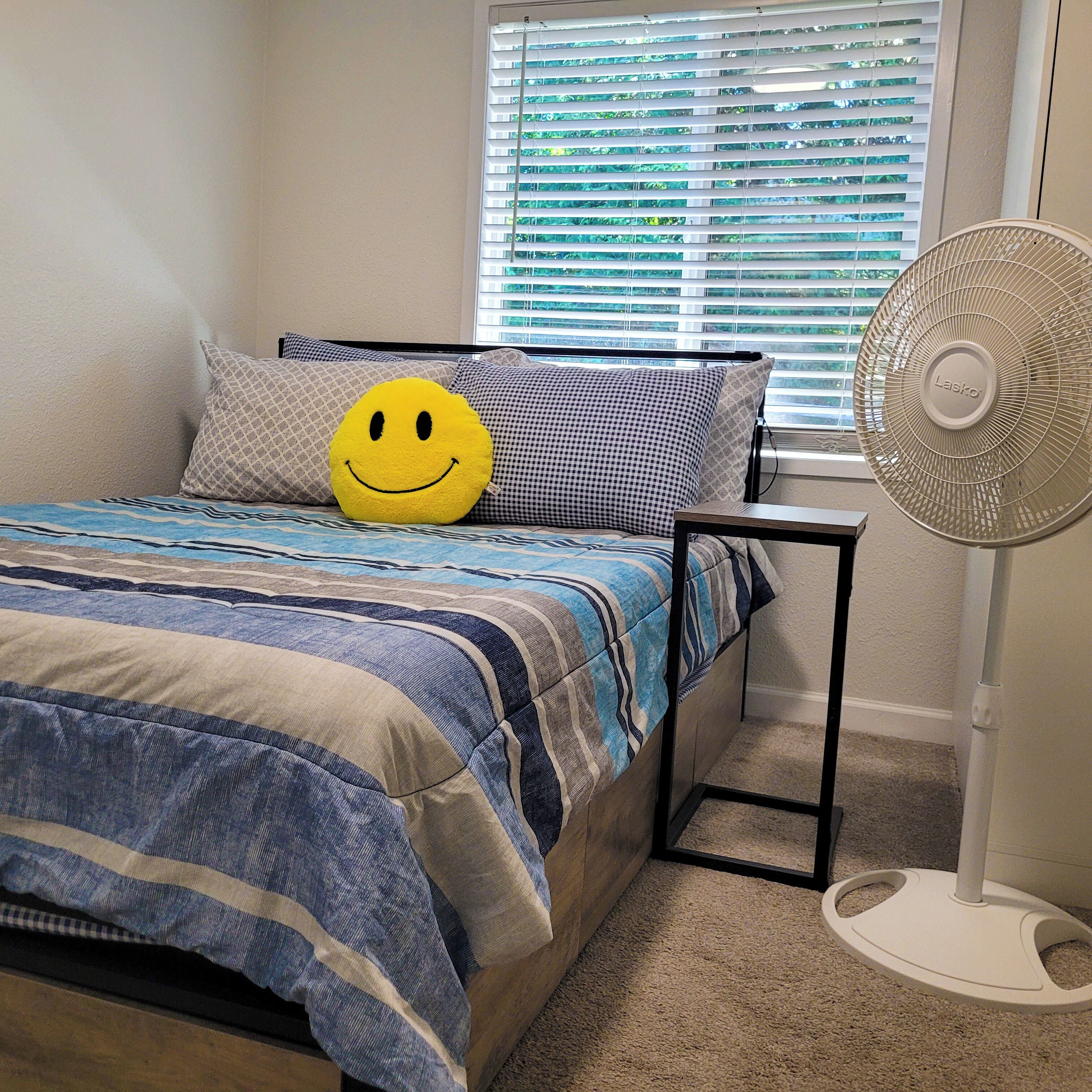 A bedroom with a bed, a smiley face pillow, a striped blanket, and a standing fan.