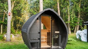 Een klein, halfopen houten blokhut met een schuin dak, een zichtbaar interieur met een houten bankje, en een deur naar binnen.