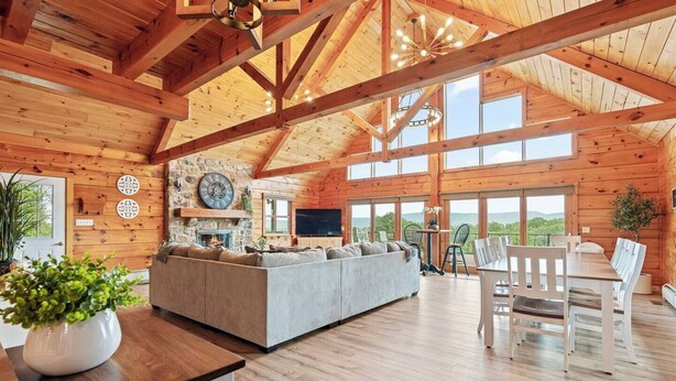 A spacious living area with a stone fireplace, wooden beams, and large windows offering a view of the outdoors. - The perfect family retreat, hot tub, fire pit, playhouse for the kids with a zip line! Plenty of fun for the young and old alike! All while enjoying the majestic views of Juniata County. (Honey Grove)