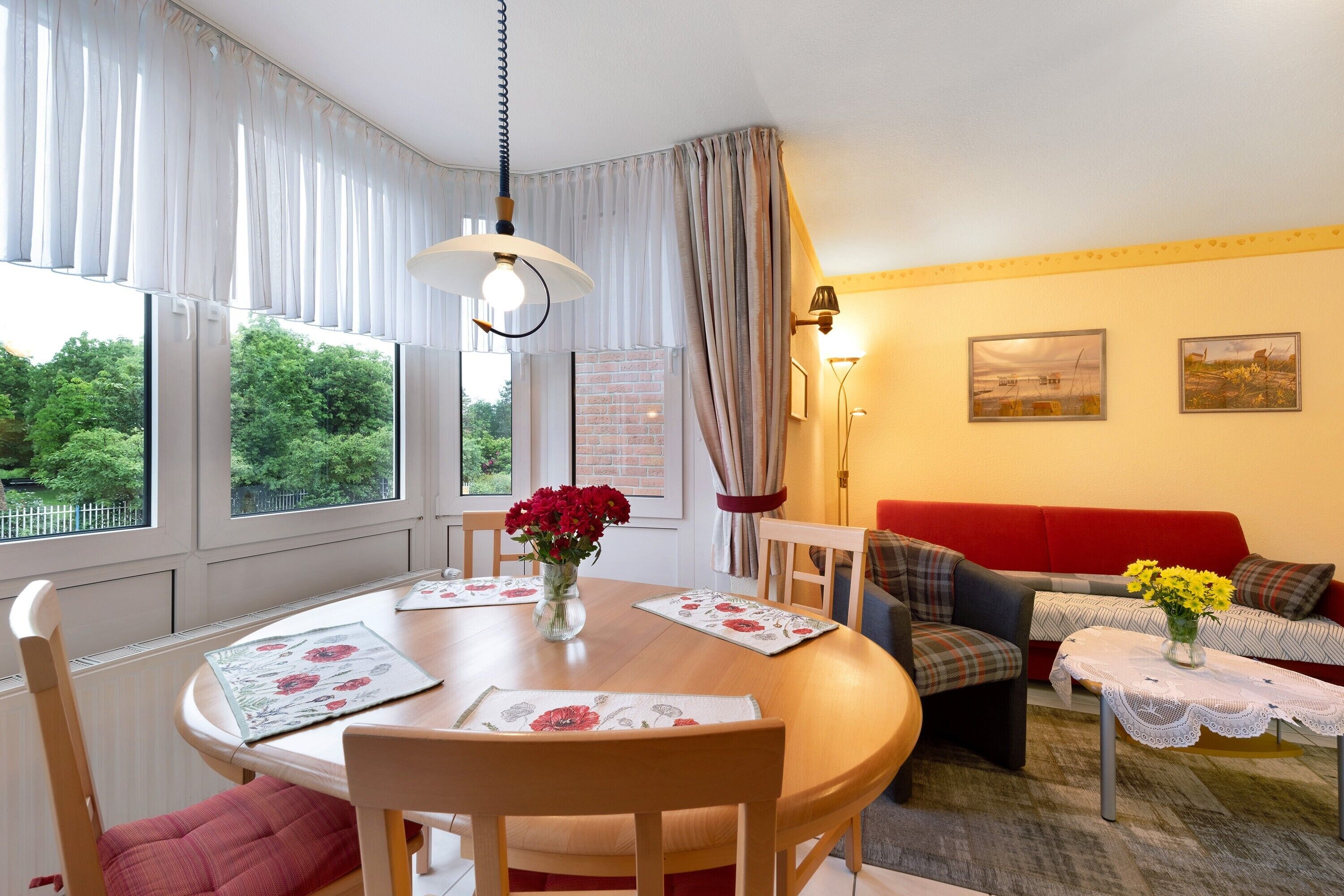 A dining area with a round table, chairs, a vase with flowers, and a red sofa in the background.