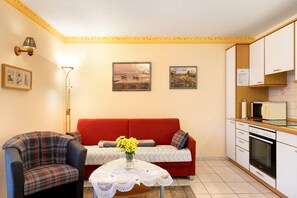 Una sala de estar con un sofá, dos sillones, una mesita con un florero, una lámpara y una zona de cocina con armarios y electrodomésticos.