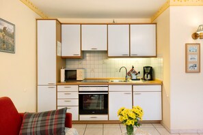 Cocina compacta con armarios blancos, microondas, cafetera y sofá rojo con cojín escocés.