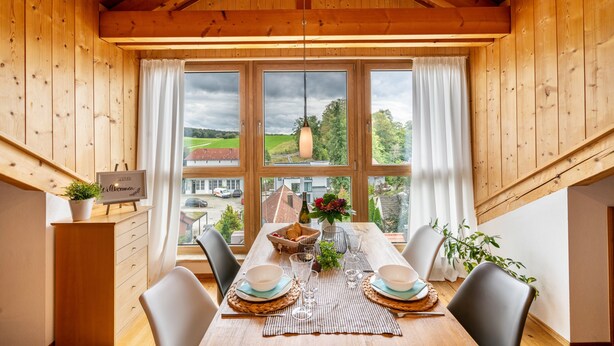 Une salle à manger en bois, avec une table dressée pour deux personnes, de grandes fenêtres et une vue sur la campagne.