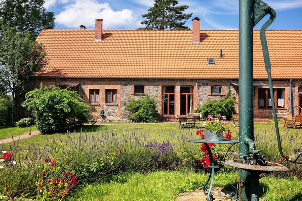 A stone building with a tiled roof, a garden with various plants, and a vintage water pump.