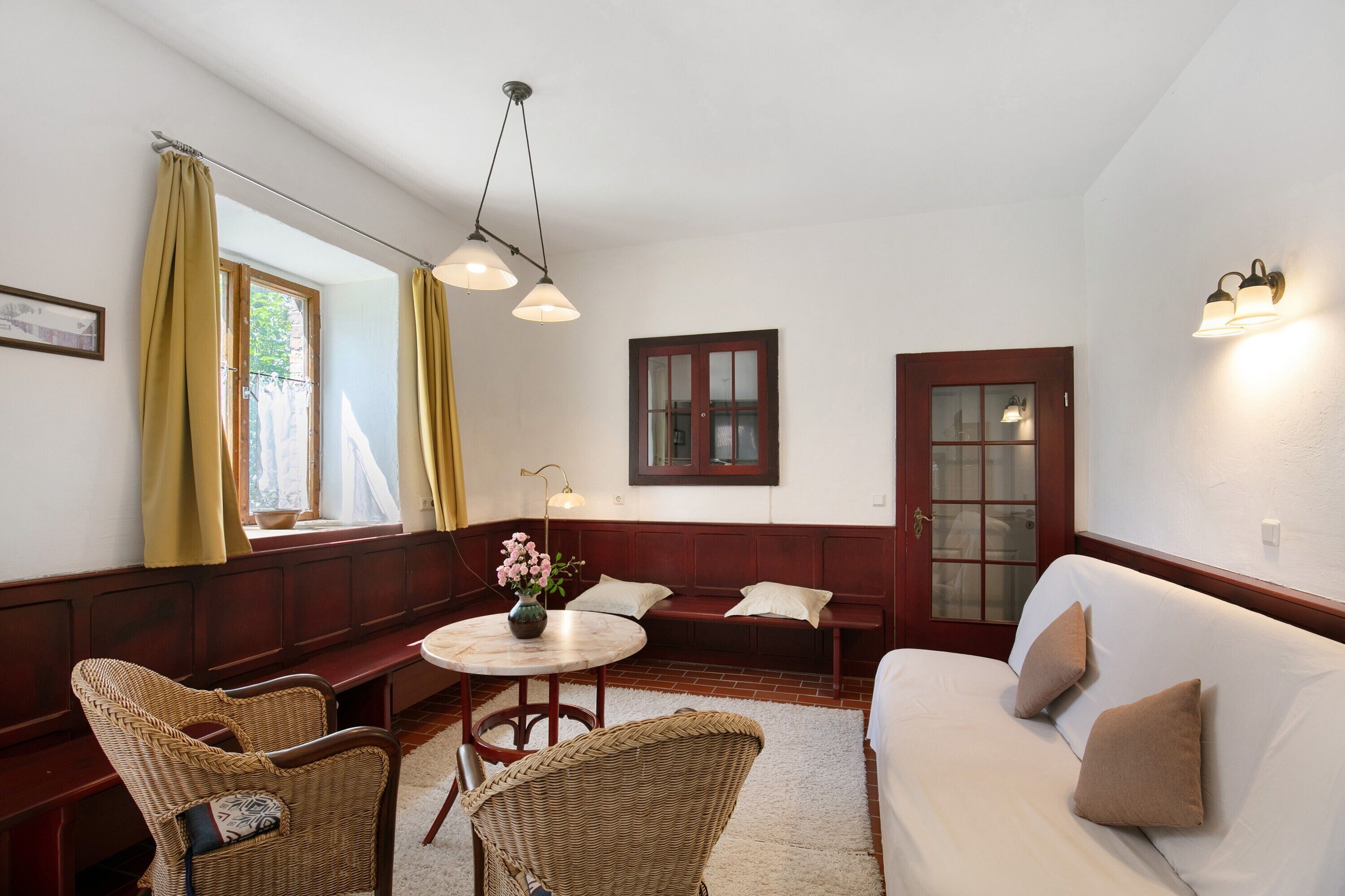 A room with a wooden bench, a round table with a vase, a sofa with cushions, and a window with curtains.