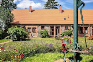 Un bâtiment en pierre avec un toit en tuiles, un jardin avec différentes espèces de plantes et une vieille pompe à eau.