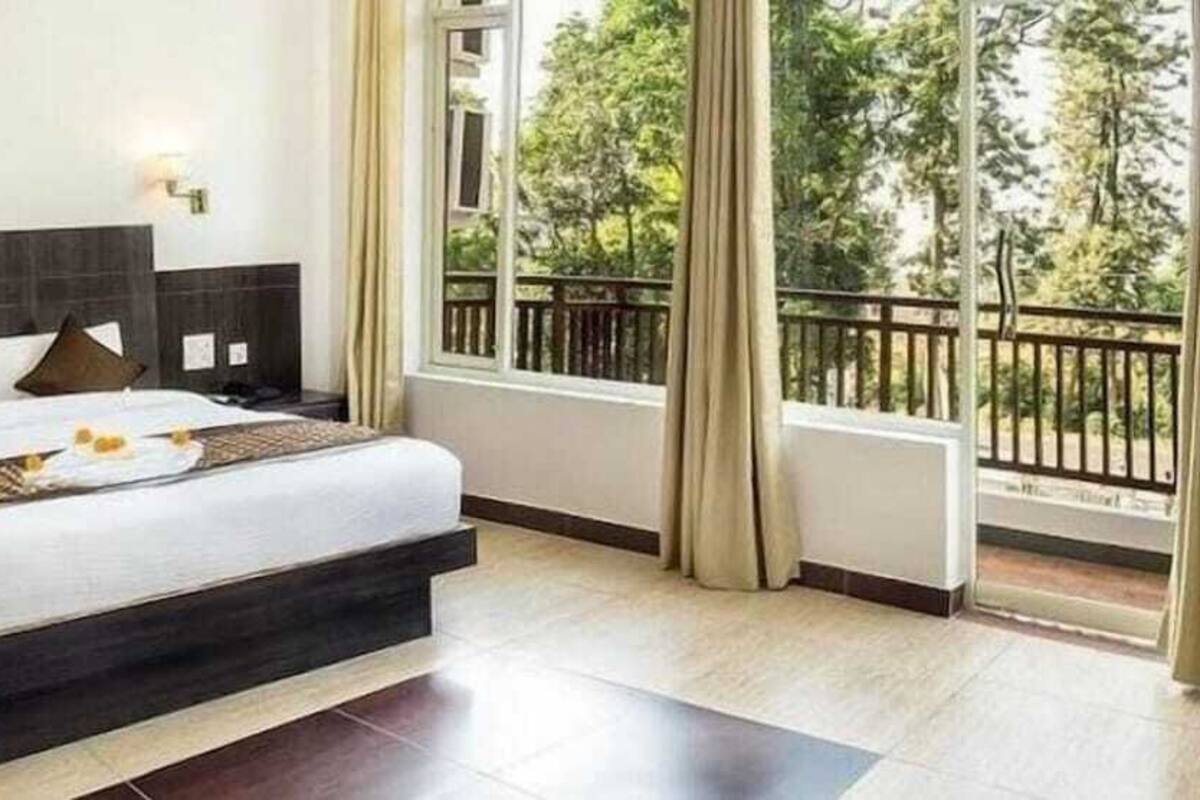 A hotel room with a large bed, a view of trees through the window, and a balcony with a railing.