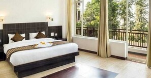 A hotel room with a large bed, a view of trees through the window, and a balcony with a railing. - Anupam Resort (Ramnagar)