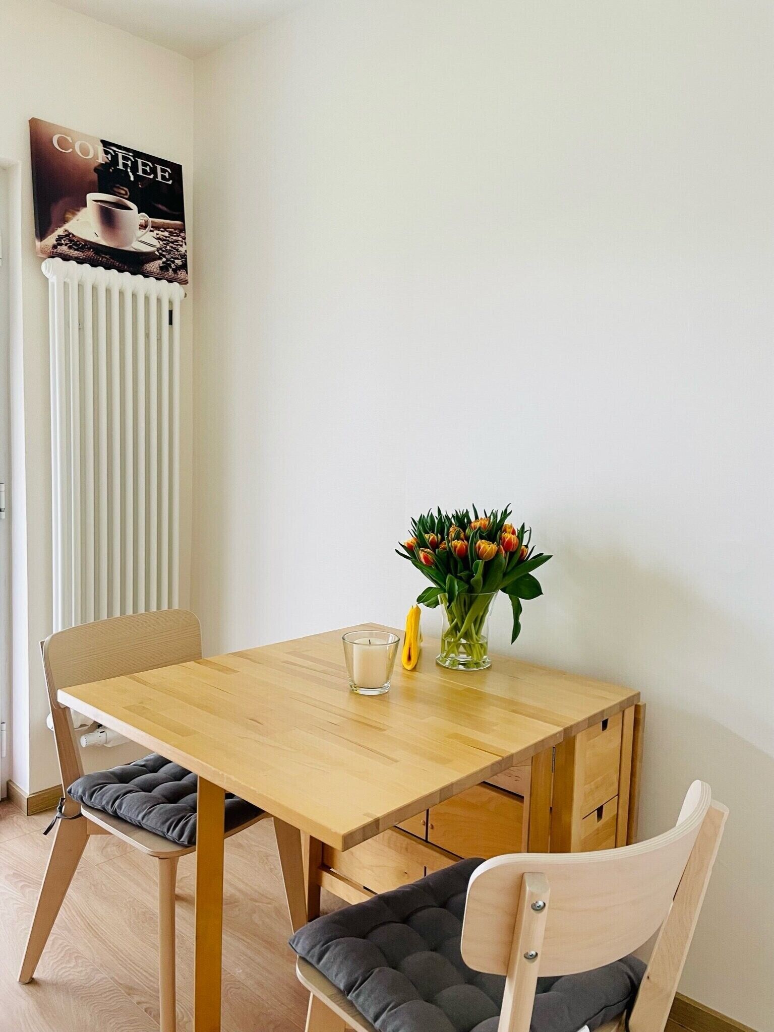 Ein Essbereich mit einem Holztisch, zwei Stühlen, einer Blumenvase, einer Kerze und einem Poster an der Wand.