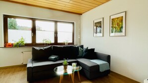Una sala de estar moderna con techo de madera, un sofá negro, una mesa de centro de vidrio y dos obras de arte enmarcadas en la pared.
