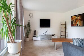 Uma sala de estar moderna com um banco de madeira, um sofá cinza, uma televisão de tela plana, um vaso de planta, uma pequena estátua e um quadro na parede.