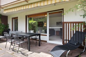 Um pátio externo com mesa e cadeiras, uma porta de correr de vidro que dá para um espaço interno e um cercado de madeira.