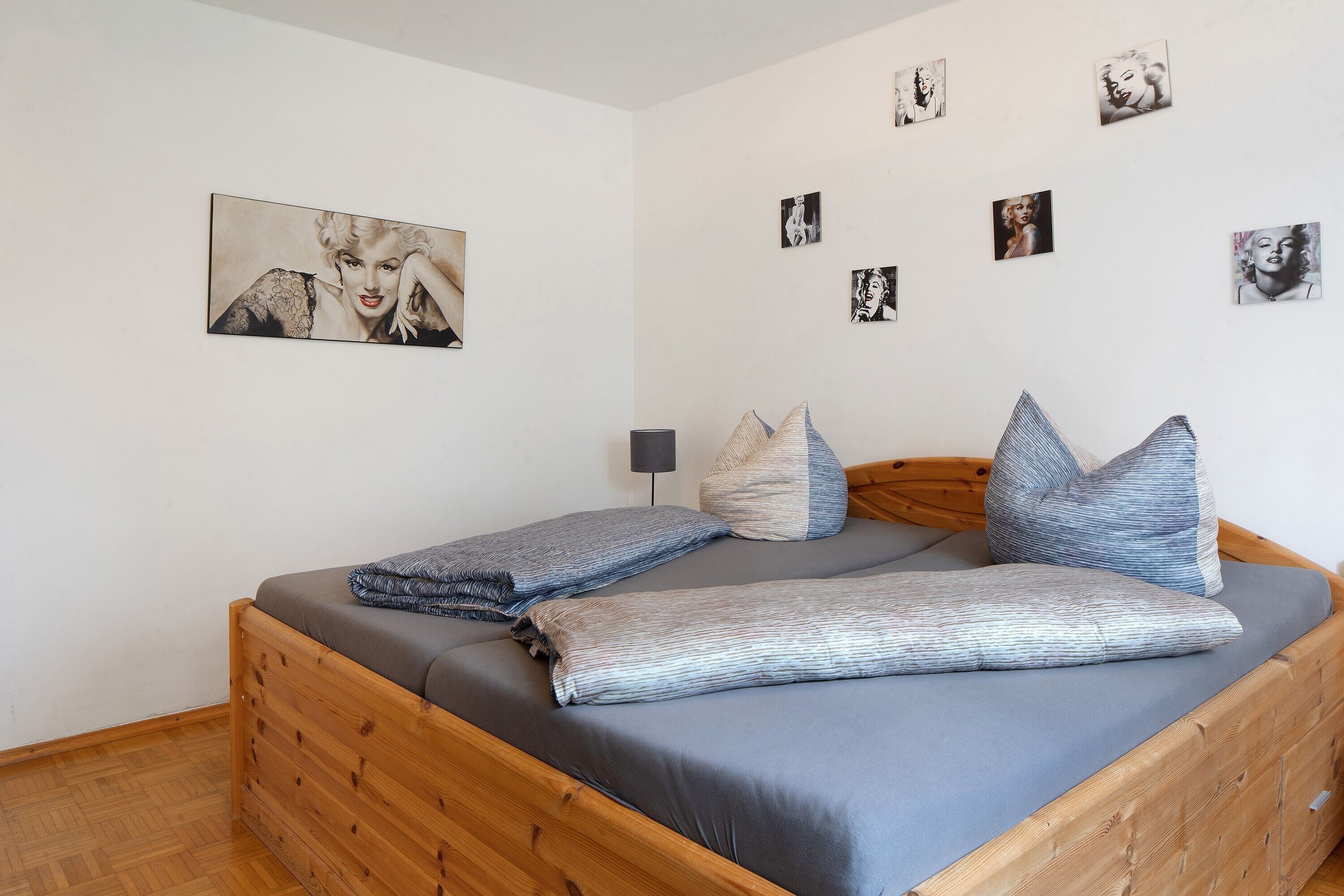 A wooden bed with grey bedding, a wall with multiple framed pictures, and a small table with a lamp.