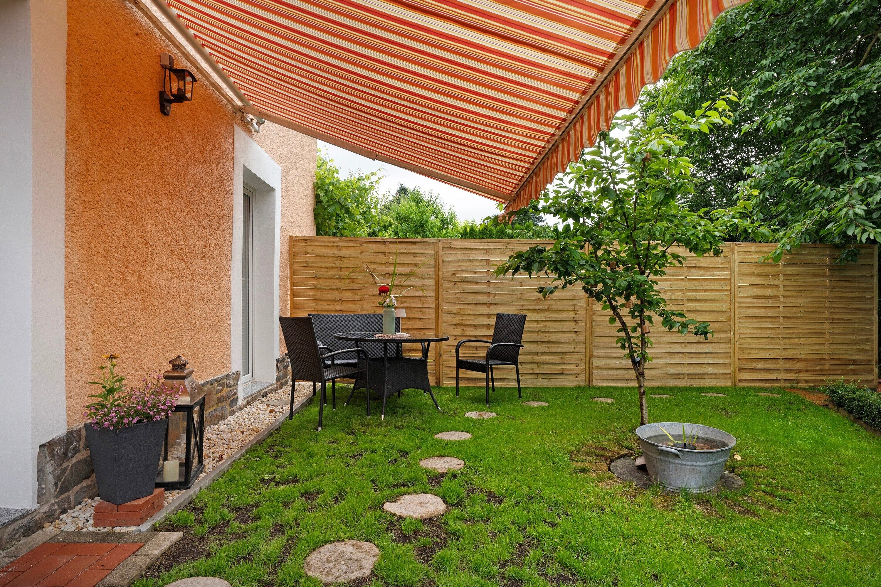 A patio with a striped awning, a wooden fence, a small tree, and outdoor furniture.