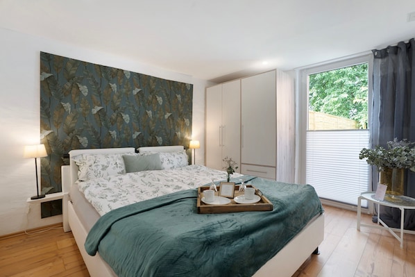 A bedroom with a bed, bedside lamps, a tray with cups and a picture frame, a window with curtains, and a wall with a leaf pattern.
