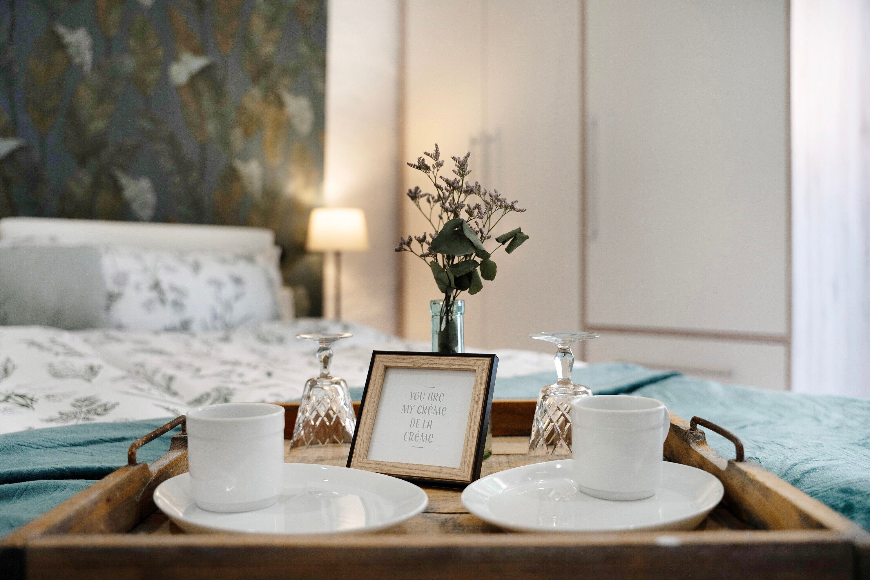 Siististi järjestetty aamiaistarjotinpohja, jossa on kaksi valkoista kuppia ja aluslautasia, kehystetty sitaatti ja maljakko kuivakukkia, kaikki puisella alustalla hotellihuoneessa.