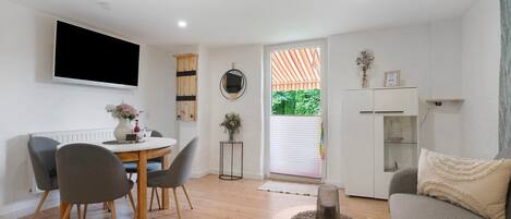 A modern living room with a dining table, chairs, a sofa, a TV, and a view through a window.