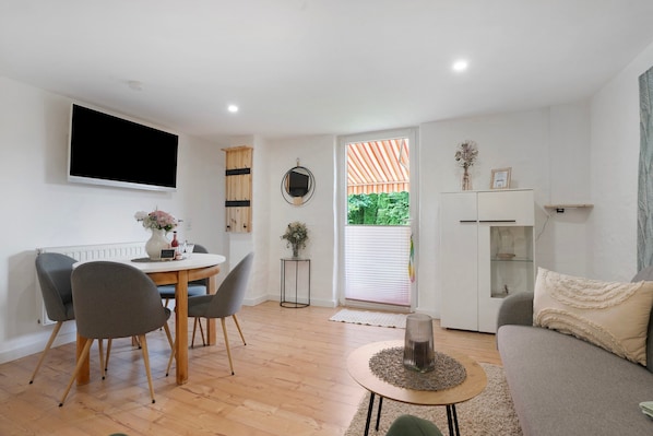 A modern living room with a dining table, chairs, a sofa, a TV, and a view through a window. - Apartment 'Grünzweig' with Private Terrace by the Water (Oelsnitz/Vogtl.)