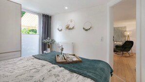 A bedroom with a bed, a bedside table with a vase, a tray with items, and a view of a garden through a window. - Apartment 'Grünzweig' with Private Terrace by the Water (Oelsnitz/Vogtl.)