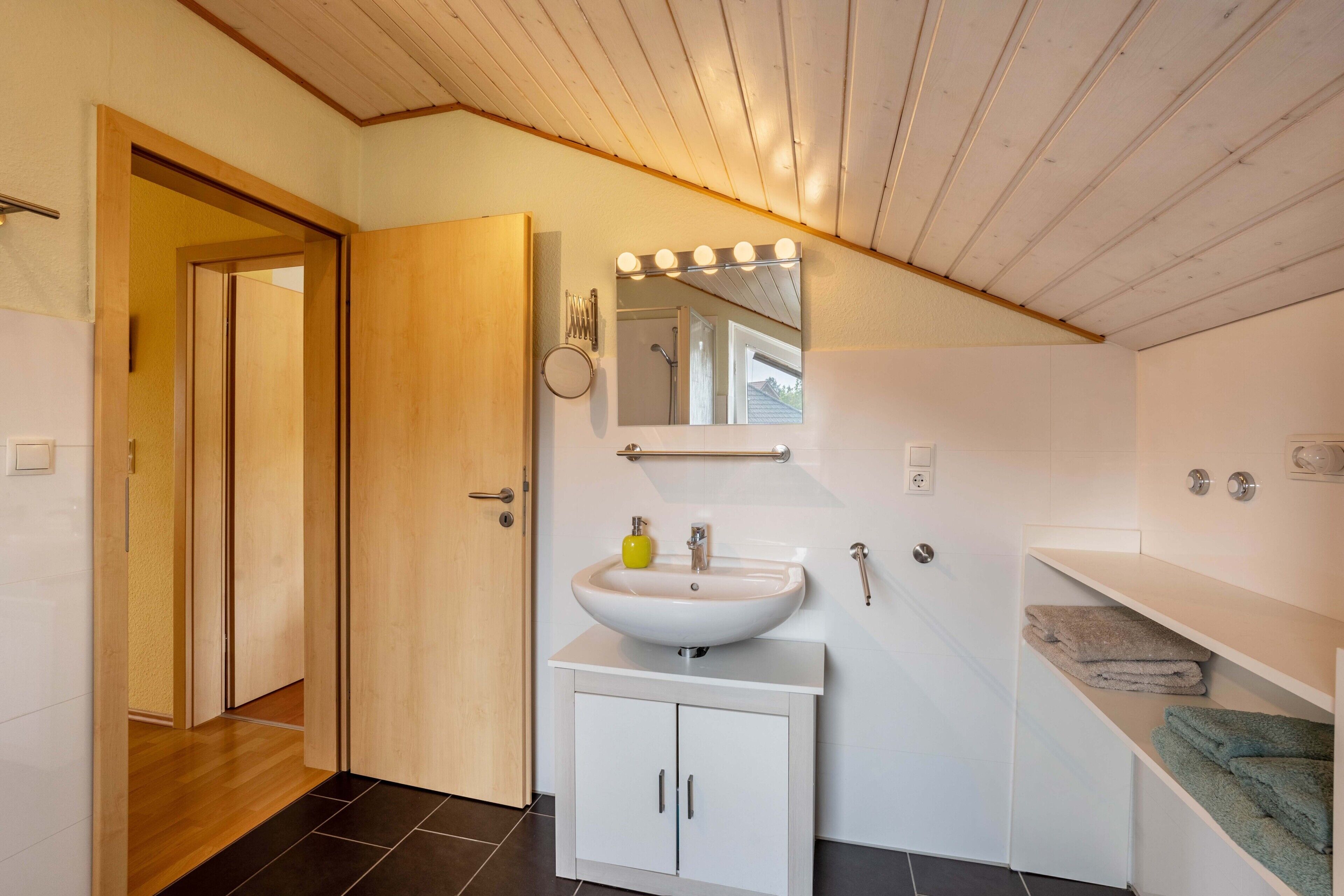 A modern bathroom with a wooden ceiling, a white sink, a mirror with lights, and a towel rack.