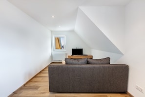 A modern loft bedroom with a built-in sofa, a television on a wooden stand, and a window with a view of a building.