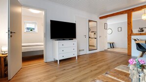 A modern room with a wooden floor, a white dresser with a flat-screen TV, a small window with a plant, and a mirror on the wall. - Apartment 'Romantische Whg In Ruhiger Lage' with Private Garden and Wi-Fi (Stelle)