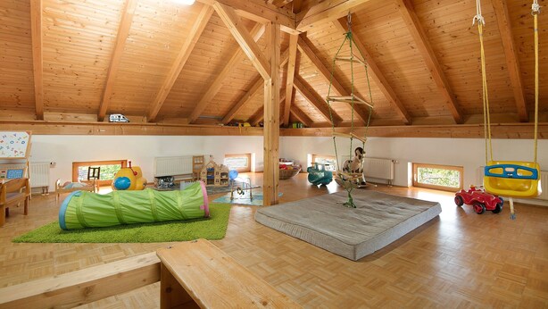 Um quarto amplo com teto de madeira alto, espaço para brincar em tons de verde, uma janela grande e piso de madeira.