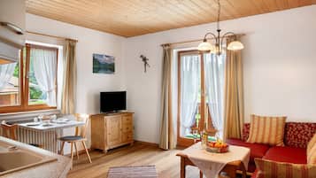 Ein gemütliches Zimmer mit Holzbalkendecke, einem Esstisch mit Stühlen, einer Couch, einem Fernseher und einem Fenster mit Vorhängen.