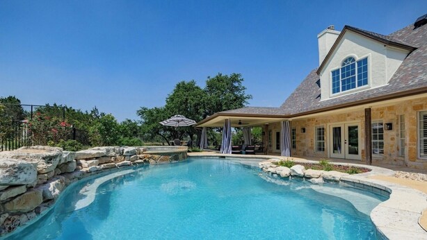 Una piscina con muros de piedra, un porche cubierto con asientos y una casa de piedra con ventanas amplias.