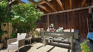 Una pérgola de madera con un banco y sillas, cerca de madera y una mesa con mantel a cuadros azules.