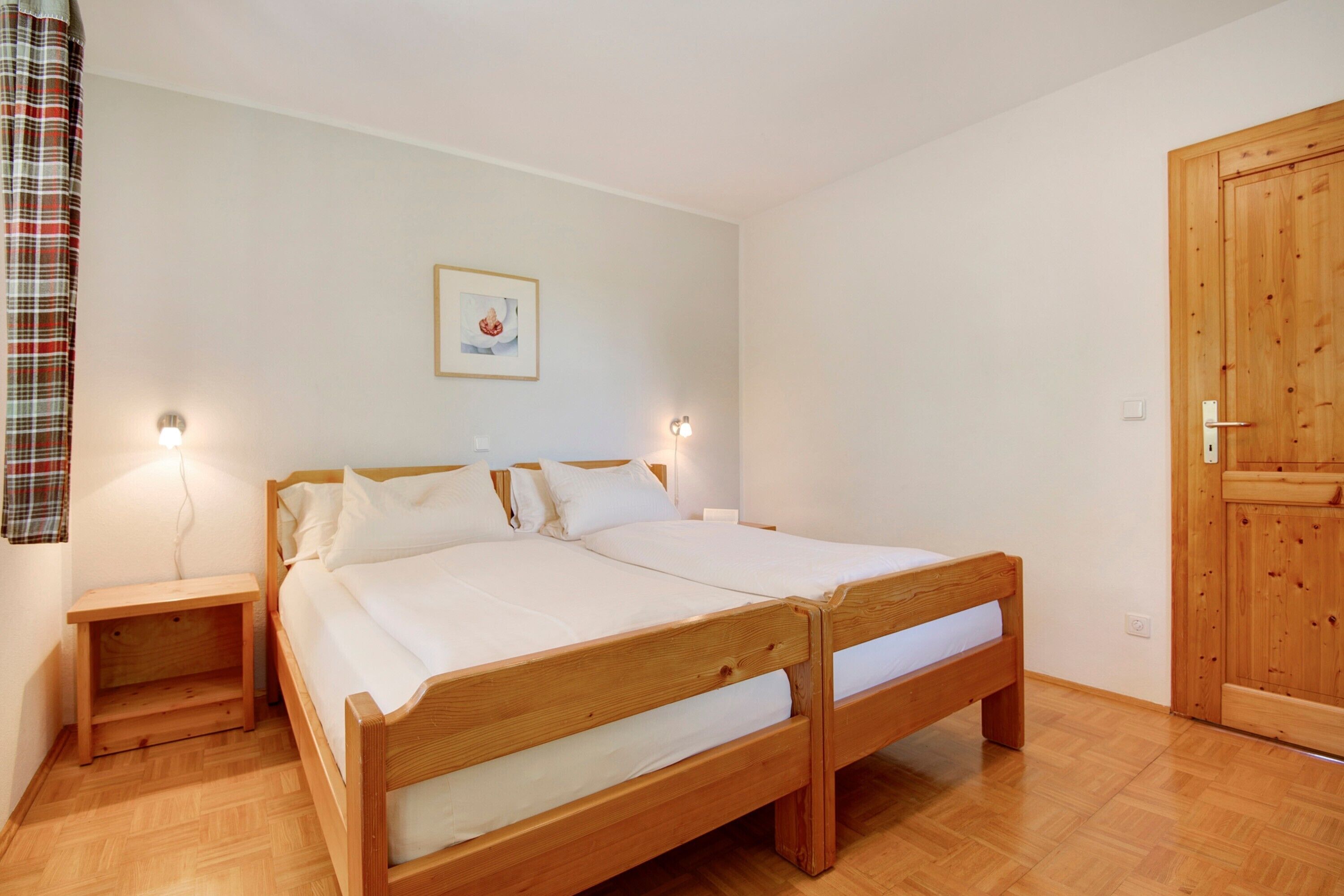 Una cama doble de madera con ropa de cama blanca, una mesita de noche, una lámpara de pared y una puerta de madera.