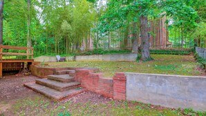 Een houten terras met trappen naar een bakstenen muur, omringd door een bebost gebied met bomen en een gebouw op de achtergrond.
