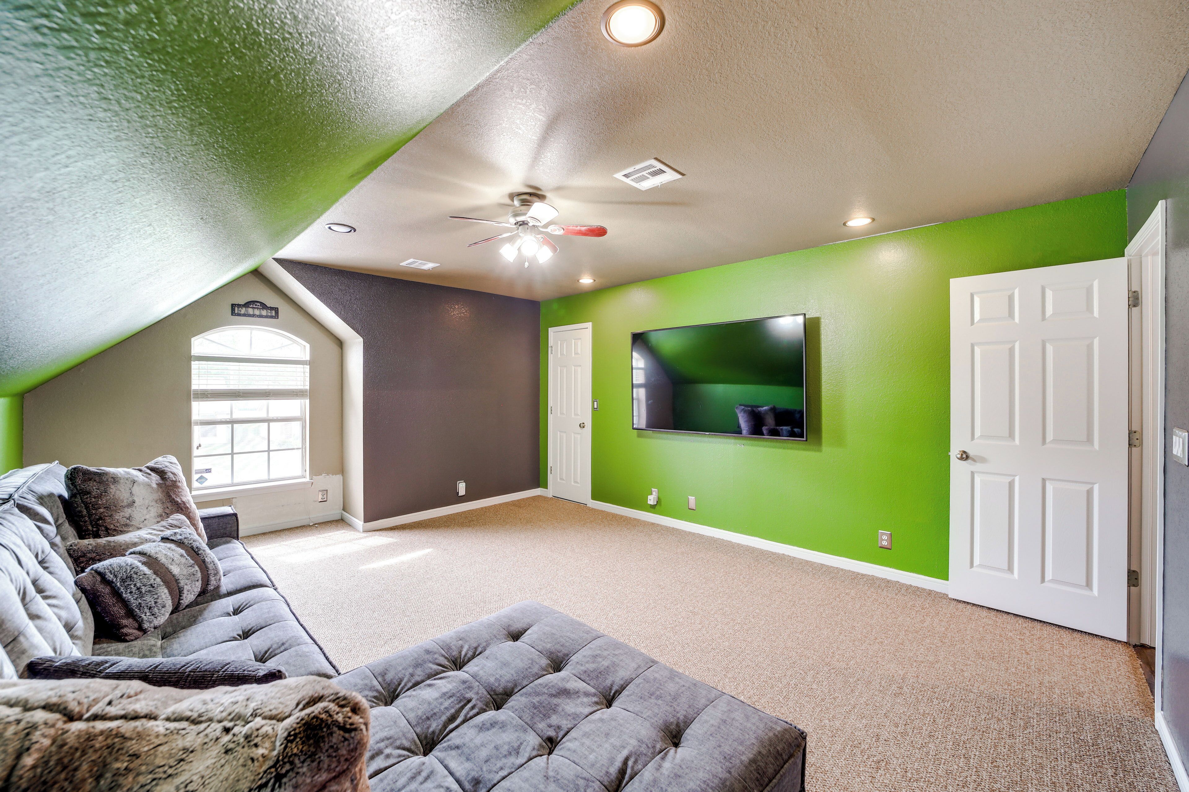 A spacious living room with a high ceiling, a ceiling fan, a large window, a grey sofa, and a green accent wall.