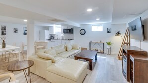 Un salon moderne avec un canapé d’angle beige, une table basse en bois et un téléviseur posé sur un meuble en bois. On aperçoit, au fond, une cuisine avec des meubles blancs et un four à micro-ondes.