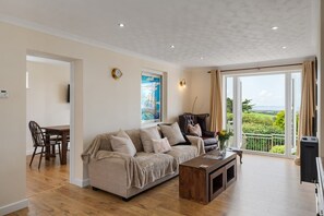 A living room with a sofa, a coffee table, a dining table with chairs, a TV, and a large window with a view. - Peaceful Home, Sea Views, Private Parking (Trewarmett)