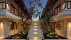 Entrada de hotel con una escalera bordeada por árboles y plantas, que conduce a un edificio con balcones.