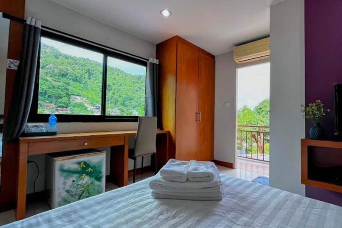 Quarto de hotel com cama, escrivaninha, televisão e varanda com vista para o verde.