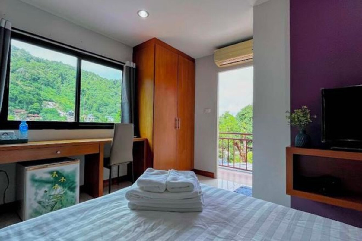 A hotel room with a bed, a desk, a wardrobe, a television, and a balcony with a view of greenery.