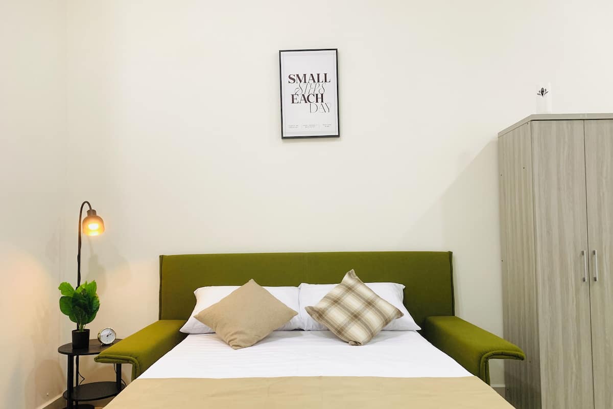 A neatly made bed with a green headboard, a bedside table with a lamp, a small potted plant, and a wardrobe.